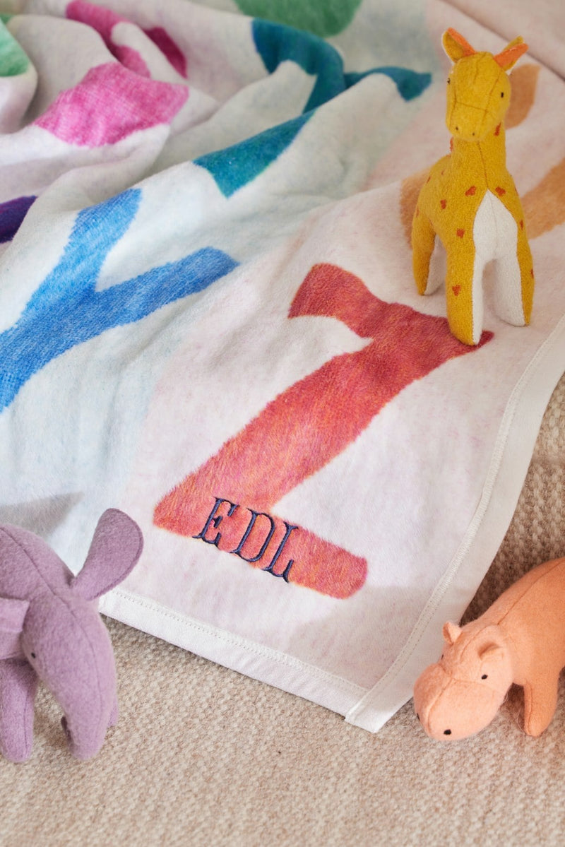 Cream blanket with colorful animal silhouettes in pink, blue, red, and purple, displayed with matching stuffed toys.