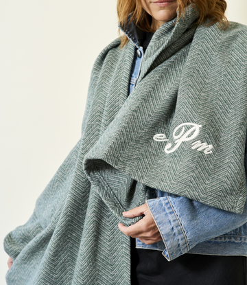 Woman wearing sage green herringbone-patterned shawl with white embroidered logo over blue shirt.8