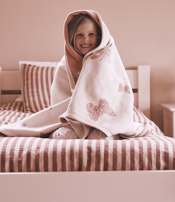 Child wrapped in cream blanket with mauve butterfly prints, sitting on striped bedding.3