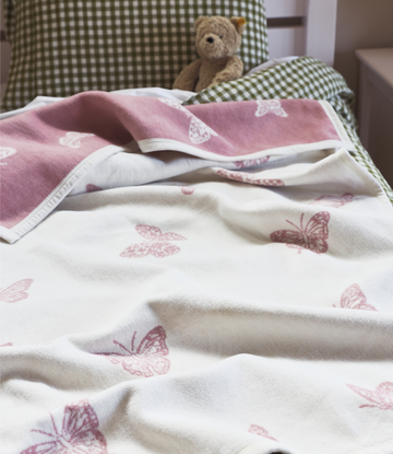 White and pink reversible blanket with butterfly print, draped on bed with green gingham pillows and teddy bear.2
