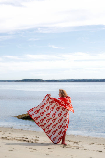 Red and white blanket with repeated lobster print worn by person on beach by water.2