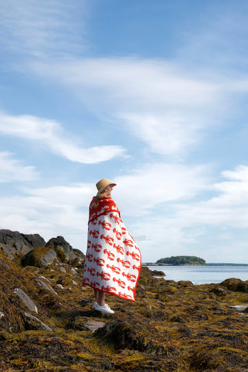 Red and white blanket with repeating lobster print worn by person standing on rocky coastal shore.3