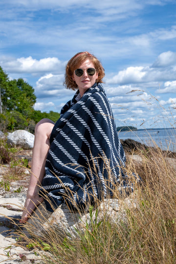 Navy blanket with white rope pattern worn by person outdoors near water and beach grass.4