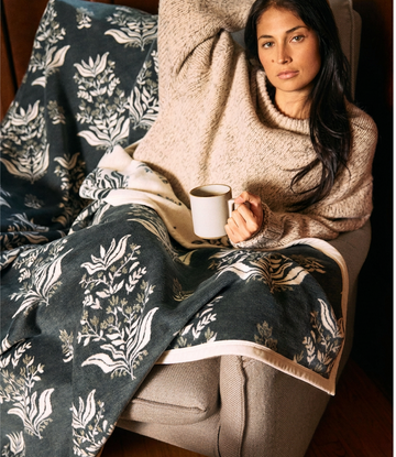 Dark blue blanket with cream botanical blockprint pattern and trim, draped over person's lap.2