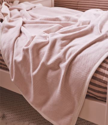 Ballet pink herringbone blanket draped on a bed with striped pillows and headboard.4