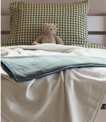 Forest green herringbone patterned blanket draped across a bed with cream pillows and stuffed bear.3