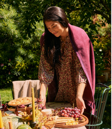 Woman draping plum-colored herringbone blanket over shoulders at outdoor picnic table with food and greenery background.2