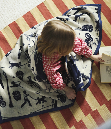 Cream blanket with navy dog and park-themed print, navy binding, laid on striped rug.3