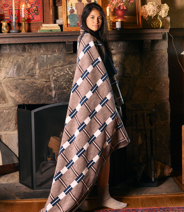 Tan blanket with navy and white geometric grid pattern draped over woman's shoulders by fireplace.3